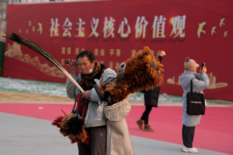 2024年1月20日,北京一名街头小贩在“社会主义核心价值观”宣传牌旁边一边听手机、一边兜售鸡毛掸子。